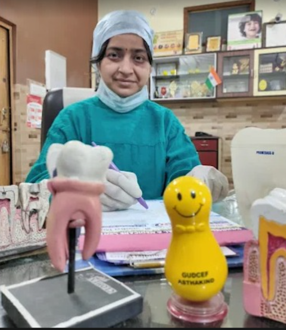 Dr. Charu Singhal in her dental clinic, wearing scrubs, with a tooth model on the desk